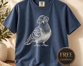 May include: A navy blue short-sleeve t-shirt featuring a white graphic illustration of a pigeon wearing round glasses. The shirt has a crew neckline and a Comfort Colors brand label on the inside collar. The fabric has a washed, vintage appearance.