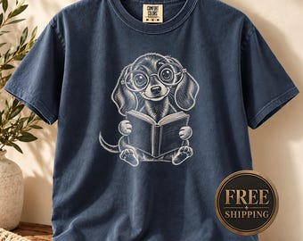 May include: A navy blue short-sleeve t-shirt featuring a white line-art graphic of a dachshund wearing round glasses and reading an open book. The shirt has a crew neckline and a Comfort Colors brand label printed on the inside of the collar.