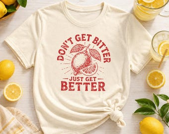 May include: A cream-colored short-sleeve t-shirt featuring a red graphic of a whole lemon and a lemon slice with the text "DON'T GET BITTER JUST GET BETTER" printed in a distressed, bold, capitalized font centered on the chest area.