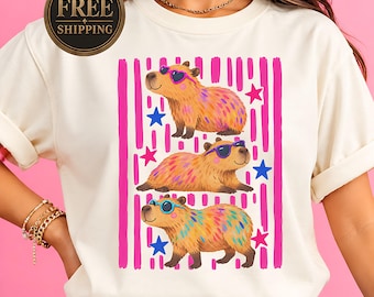 May include: A cream-colored t-shirt featuring a graphic of three capybaras wearing colorful sunglasses. The design includes vertical pink stripes and scattered blue and pink stars. The capybaras have multi-colored fur patterns in orange, yellow, and blue.
