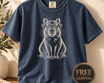 May include: A navy blue short-sleeve t-shirt featuring a white line-art graphic of a capybara wearing round glasses. The shirt has a crew neckline and a Comfort Colors brand label on the inside collar. The fabric has a washed, vintage appearance.