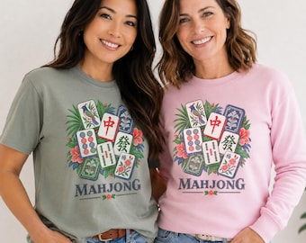May include: A sage green short-sleeve t-shirt and a light pink long-sleeve sweatshirt, both featuring a colorful graphic print of Mahjong tiles surrounded by tropical leaves and hibiscus flowers, with the word MAHJONG printed in dark blue lettering.