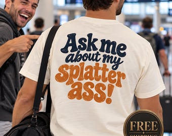 May include: An off-white short-sleeve t-shirt featuring the text "Ask me about my splatter ass!" printed on the back. The words "Ask me about my" are in dark blue, while "splatter ass!" is in orange with a distressed, textured appearance.
