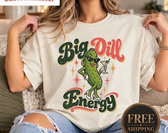 May include: A cream-colored t-shirt featuring a graphic of a green pickle wearing sunglasses, gold hoop earrings, and white high-heeled shoes while holding a martini glass. The text Big Dill Energy is printed in large, stylized green and red lettering.