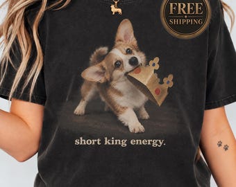 May include: A charcoal gray t-shirt featuring a graphic of a brown and white Corgi puppy holding a gold paper crown in its mouth. Below the puppy, the text reads "short king energy." The shirt has a crew neckline and short sleeves.