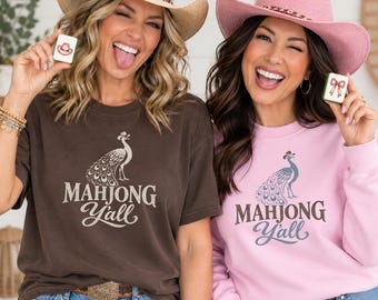 May include: A dark brown short-sleeve t-shirt and a light pink long-sleeve sweatshirt, both featuring a graphic of a peacock and the text "MAHJONG Y'ALL". The t-shirt is paired with a straw cowboy hat, and the sweatshirt with a pink cowboy hat.