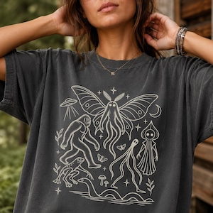 May include: A charcoal gray short-sleeve t-shirt featuring a cream-colored graphic print of a moth, a sasquatch, a frog, a snake, a hooded figure, a flying saucer, a crescent moon, and small mushrooms arranged in a central design.