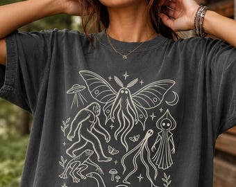May include: A charcoal gray short-sleeve t-shirt featuring a cream-colored graphic print of a moth, a sasquatch, a frog, a snake, a hooded figure, a flying saucer, a crescent moon, and small mushrooms arranged in a central design.