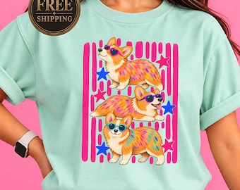 May include: A light mint green t-shirt featuring a graphic print of three Corgi dogs wearing colorful heart-shaped and round sunglasses. The design includes vertical pink brushstroke stripes and scattered blue and pink stars across the center.