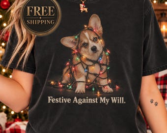 May include: A black short-sleeve t-shirt featuring a graphic of a brown and white Corgi puppy tangled in colorful Christmas string lights. Below the puppy, the text reads "Festive Against My Will." The shirt has a crew neckline and a relaxed fit.