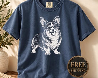 May include: A navy blue short-sleeve t-shirt featuring a white graphic illustration of a Corgi dog wearing round glasses. The shirt has a crew neckline and a Comfort Colors brand label on the inside collar, with the text M-M-M visible on the tag.