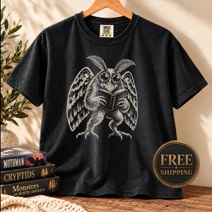 May include: A black short-sleeve t-shirt featuring a white graphic illustration of a moth-like creature wearing round glasses and reading an open book. The shirt has a crew neckline and a Comfort Colors brand label printed on the inside collar.