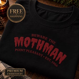 May include: A black crewneck sweatshirt featuring red embroidered text that reads BEWARE THE MOTHMAN, POINT PLEASANT, and EST. 1966. The word MOTHMAN is displayed in a large, textured, distressed font across the center of the chest.