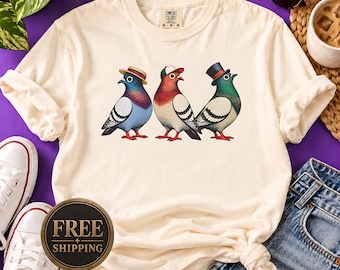 May include: A cream-colored short-sleeve t-shirt featuring a graphic of three pigeons standing in a row. The first pigeon wears a straw boater hat, the second a baseball cap, and the third a black top hat. Each bird has distinct plumage colors.