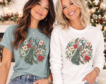 May include: A sage green t-shirt and a white long-sleeve sweatshirt, each featuring a circular graphic of a peacock surrounded by poinsettias, pine branches, and four mahjong tiles with Chinese characters, worn with blue denim jeans.