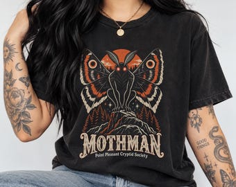 May include: A black t-shirt featuring a graphic of a winged creature with glowing red eyes perched on a rocky mountain peak. The design includes orange and white accents, a forest silhouette, and the text MOTHMAN Point Pleasant Cryptid Society.