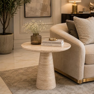 Puede incluir: Una mesa auxiliar redonda de travertino beige con base cónica. Sobre la mesa hay un jarrón de vidrio oscuro estriado con flores blancas de paniculata, un pequeño cuenco dorado y dos libros apilados titulados Kerry Joyce y The Intangible.
