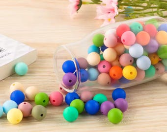 May include: A collection of small, round, matte-finish silicone beads in a wide variety of vibrant colors, including blue, green, pink, yellow, orange, and purple, spilling out from a clear glass bottle onto a light-colored wooden surface.