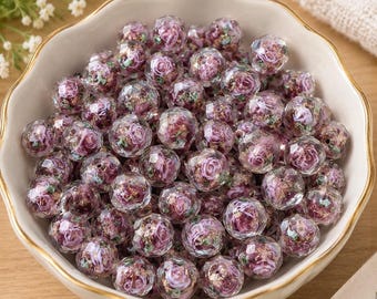 May include: A collection of round, faceted clear beads contained within a scalloped white bowl. Each bead features an internal design of a purple rose surrounded by gold and green glitter accents, creating a layered, three-dimensional appearance.