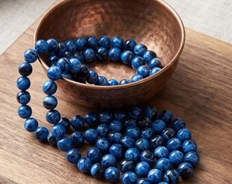 May include: A strand of round, polished blue beads with dark marbled patterns, partially coiled inside a small, round, hammered copper bowl. The beads feature varying shades of deep and royal blue with black veining throughout the surface.