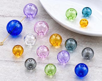 May include: A collection of round, translucent glass beads in various colors including blue, purple, yellow, green, clear, and grey. One blue bead is threaded onto a thin gold-toned metal wire. The beads are scattered, with some resting inside a white dish.