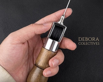 May include: A sewing awl featuring a wooden handle, a silver metal body, and a sharp needle. The tool includes an internal spool of black thread visible through a clear window. The text DEBORA COLECTIVES and Premium Sewing Awl are present.