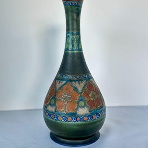 Może przedstawiać: Wysoki ceramiczny wazon z wąską szyjką i pękatą podstawą, pokryty ciemnozielonym szkliwem. Korpus ozdobiony jest pasem pomarańczowych i czerwonych motywów kwiatowych, podkreślonych niebiesko-białymi wzorami geometrycznymi oraz drobnymi kolistymi detalami.