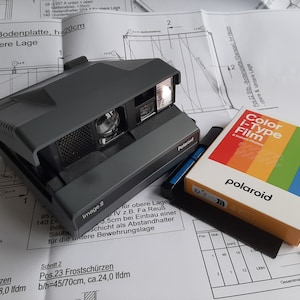May include: A grey Polaroid instant camera with a flash, beside a box of Color i-Type film. The film box has a rainbow design. A black squeegee and a blue pen are also present. The items are on a blueprint.