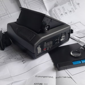 May include: A black and grey electronic device with an open top, a lens, and various buttons and ports. A black accessory and a small black case with a silver ring and blue batteries are also present. The items rest on a blueprint.