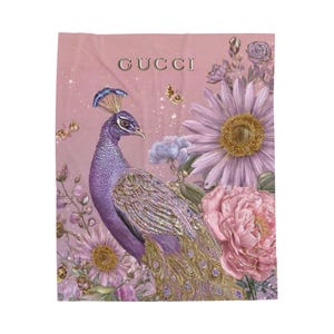 Può includere: Un tessuto rettangolare con sfondo rosa polvere e la scritta GUCCI stampata in oro. Il design mostra un pavone viola con piumaggio dorato circondato da grandi margherite color lavanda, peonie rosa e piccole farfalle dorate.
