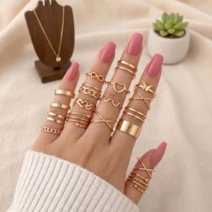 Peut inclure: Une collection de bagues dorées portées sur les doigts, présentant divers motifs dont un symbole infini, un cœur, un papillon, des maillons de chaîne, des bandes ondulées et des anneaux lisses. Les bagues sont portées sur plusieurs phalanges.