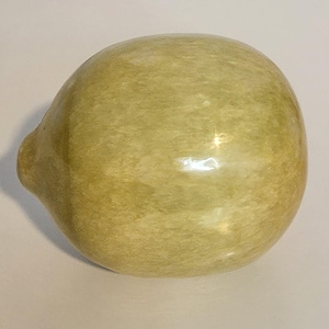 May include: A smooth, rounded ceramic object in a pale olive-green hue with a mottled, variegated surface texture. The item features a small, protruding nub on the left side and a glossy finish that reflects light across its curved, bulbous form.