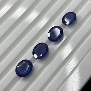 May include: Four oval-cut, deep blue sapphires arranged in a diagonal line. Each gemstone features a faceted surface that reflects light, showcasing a translucent, dark blue color profile. The stones are positioned on a white, ribbed surface.