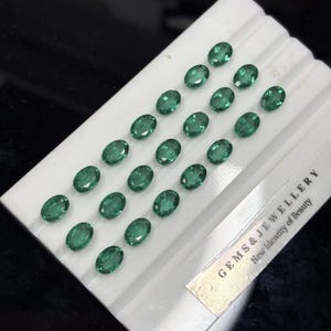 May include: A collection of twenty oval-cut green gemstones arranged in rows on a white display stand. The stand features the text GEMS & JEWELLERY New Identity of Beauty printed in black on a silver-toned label at the bottom right corner.