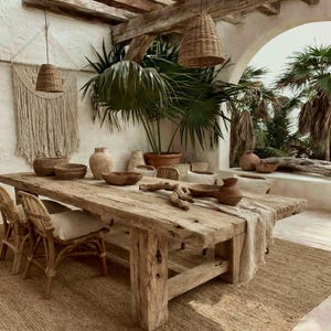 May include: A rustic dining area featuring a long, weathered wooden table with a light-colored runner, surrounded by woven rattan chairs. The space includes several ceramic bowls, a macrame wall hanging, and two hanging wicker pendant lights.