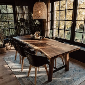 Pode incluir: Uma mesa de jantar retangular de madeira com borda natural e acabamento de veios visíveis, apoiada por pernas de madeira robustas. A mesa é cercada por quatro cadeiras pretas com pernas de madeira clara e iluminada por uma luminária pendente trançada.