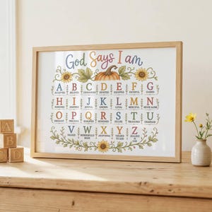 May include: A framed wall art print titled God Says I Am featuring an alphabet chart with colorful letters and corresponding words. The design includes illustrations of sunflowers and a pumpkin, all enclosed in a light wood frame on a wooden surface.