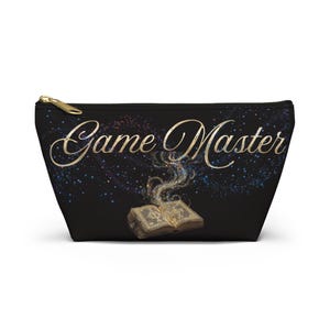 May include: A black zippered pouch featuring the gold text Game Master in a cursive font. Below the text is an illustration of an open, ornate book with glowing, swirling magical energy rising from its pages against a dark, starry background.