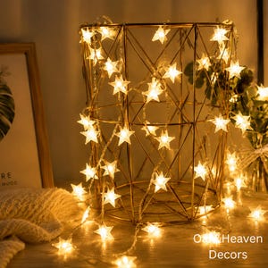 May include: A string of warm white LED star-shaped lights draped over a gold-toned geometric wire basket. The lights emit a soft glow, illuminating the metal structure. The text "Oat & Heaven Decors" is visible in the bottom right corner.