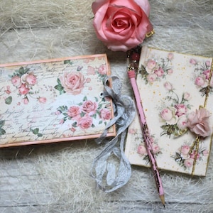 Op de afbeelding: Een rechthoekig notitieboek en een verticaal dagboek met een crèmekleurige achtergrond, roze rozenpatronen en cursieve tekst. Een roze roos ligt boven de items en een roze potlood met een decoratieve bovenkant rust op het dagboek.