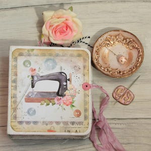 May include: A square notebook with a vintage-style sewing machine illustration, a pink fabric rose, a small ornate gold-toned dish containing a needle, and a pink ribbon closure. The notebook cover features floral accents and button graphics.