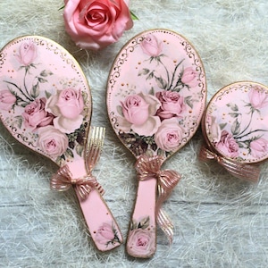 Può includere: Due specchi a mano ovali e uno specchio compatto rotondo, tutti caratterizzati da uno sfondo rosa chiaro con un dettagliato motivo floreale di rose, bordi decorativi dorati e un nastro metallico color oro rosa legato ai manici.
