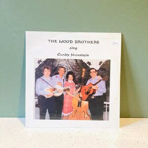 May include: A white record album cover with a photo of The Wood Brothers band. The band is playing musical instruments and smiling. The album title is "Candy Mountain" and the text "The Wood Brothers sing" is printed above the title.
