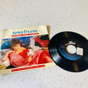 May include: A vintage vinyl record and album cover featuring Gloria Estefan and Miami Sound Machine. The album cover has a photo of Gloria in a red outfit, with the text "Rhythm Is Gonna Get You". The record is black.