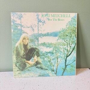 May include: Album cover for Joni Mitchell's "For The Roses." The album art features a woman in a green velvet outfit sitting near a body of water. The album title and artist name are in green text.