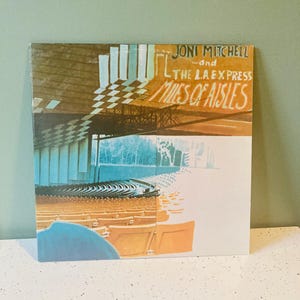 May include: Album cover for Joni Mitchell and The L.A. Express's "Miles of Aisles." The cover art features a split design with a checkerboard pattern, architectural elements, and a concert hall scene in shades of brown, blue, and orange.