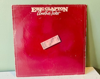 Puede incluir: Portada de álbum de vinilo rojo vintage para "Another Ticket" de Eric Clapton. El título del álbum y el nombre del artista están en texto blanco. Un pequeño gráfico de boleto aparece en la parte inferior de la portada.