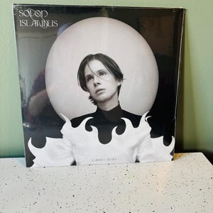 May include: A vinyl record album featuring a black and white image of a person with long hair against a white background. The album cover has the text "Sodon Islandus" at the top and "Gabriel Olars" at the bottom.