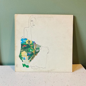 May include: Vintage album cover featuring a line drawing of a woman with a colorful landscape design. The landscape includes a yellow house, trees, and a red car. The album cover is on a white surface.