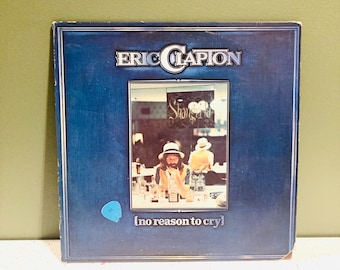 Puede incluir: Una portada de disco de vinilo azul con el texto "Eric Clapton" en letras blancas. La portada del álbum presenta una foto de Eric Clapton sentado en un bar con una bebida frente a él. El texto "[no reason to cry]" está impreso en letras blancas en la parte inferior de la portada del álbum.